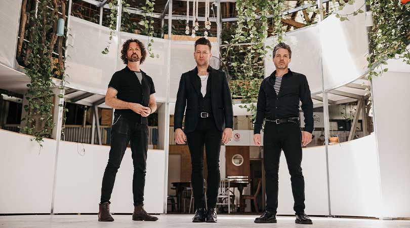 Eskimo Joe band members. 3 men wearing black standing in front of a white curved wall with green shrubbery hanging down