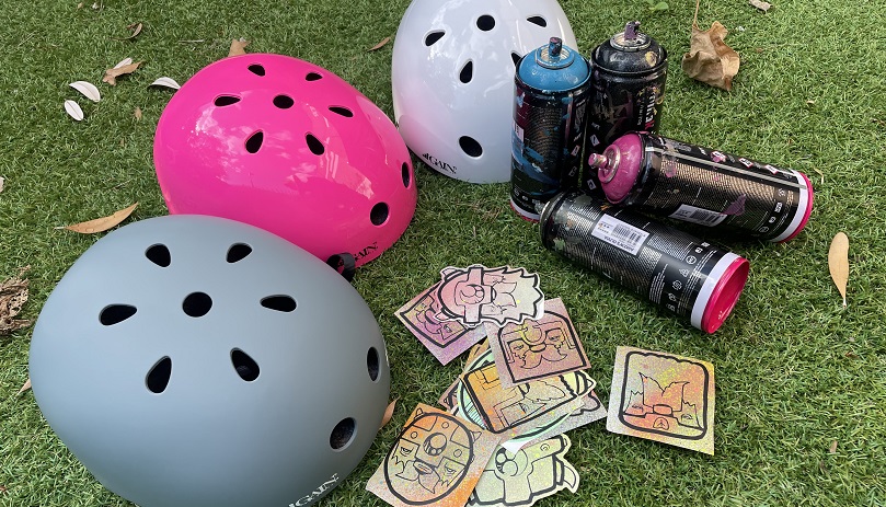 Three helmets (one grey, one pink and one white) arranging on the grass next to some Viking stickers and spray-paint cans.