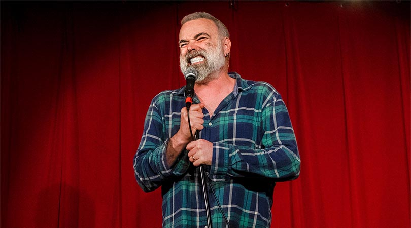 bearded man in blue flannelette shirt holding microphone and gritting teeth with red curtain background